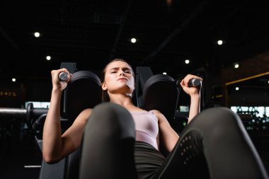 young sportswoman exercising on arm extension machine in sports center on blurred foreground clipart