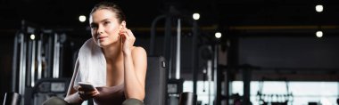 Spor kadını elinde cep telefonu tutarken gülümsüyor ve spor salonunda kablosuz kulaklıkla müzik dinliyor.