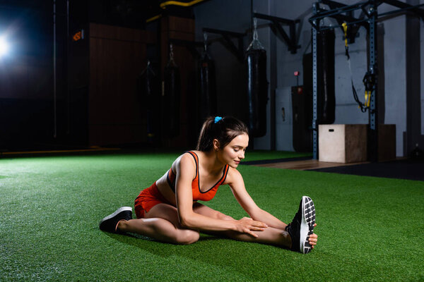 athletic sportswoman touching sneaker while doing stretching exercise in gym