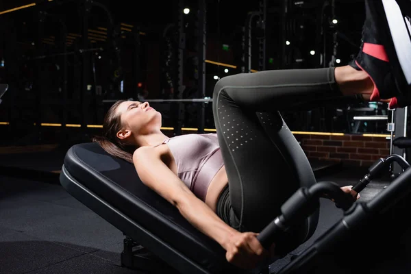 athletic woman with closed eyes exercising on leg press machine on blurred foreground