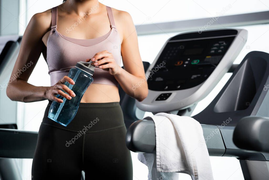 cropped view of sportswoman in top holding sports bottle while standing on treadmill