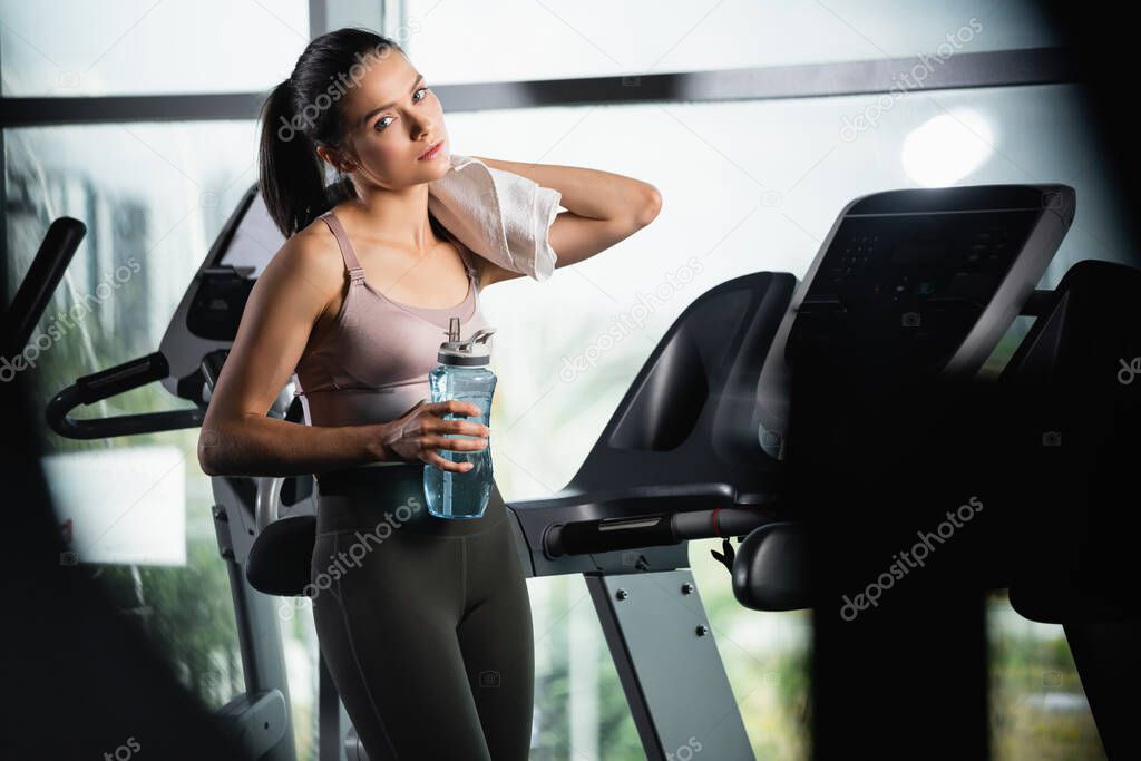 tired sportswoman standing on treadmill, wiping neck with towel and holding sports bottle on blurred foreground