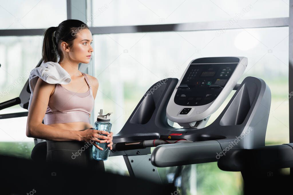 young sportswoman looking away while standing on treadmill with sports bottle on blurred foreground