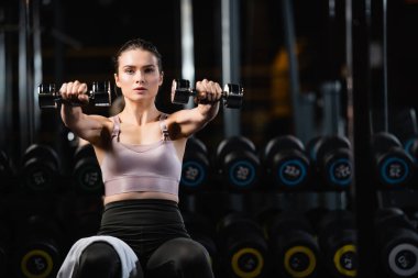 Sportif kadın oturma pozisyonunda duran dambıllarla çalışırken kameraya bakıyor.