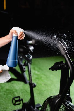 partial view of charwoman spraying cleanser on sports equipment in gym clipart