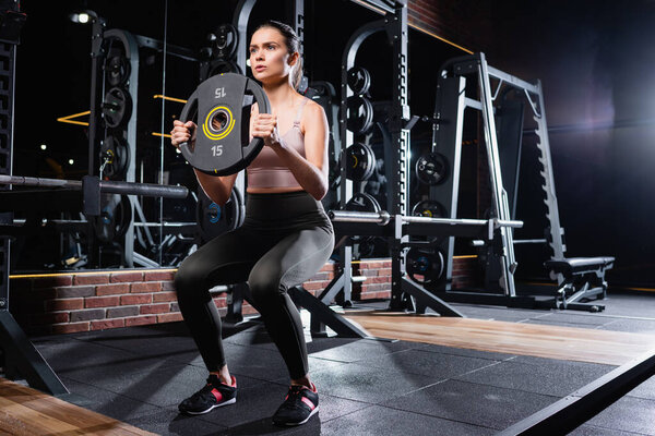 молодая спортсменка в леггинсах и кроссовках делает приседания с диском веса в тренажерном зале