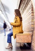 side view of woman in stylish autumn clothes holding newspaper while sitting on metro platform bench