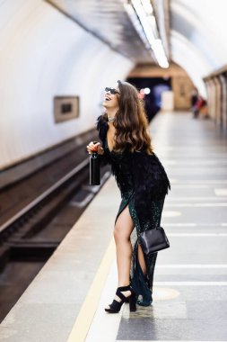 Siyah lurex elbiseli, güneş gözlüklü çekici kadın metro istasyonunda şarap şişesi tutarken gülüyor.