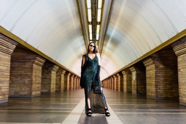elegant woman in long black dress and sunglasses posing at underground station clipart