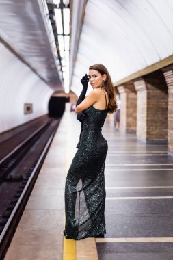 Uzun siyah elbiseli şehvetli kadın yeraltı platformunda dururken kameraya bakıyor.