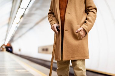Bastonlu, sonbahar montu giymiş, metro platformunda duran yaşlı bir adamın kırpık manzarası.