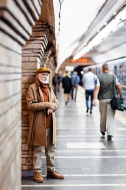 Sonbahar kıyafetli yaşlı bir adam insanların yanında duruyor ve bulanık ön planda metro platformunda eğitim görüyor.
