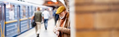 Sonbahar kıyafetli son sınıf öğrencisi metro peronunda duruyor. Önü bulanık tren ve yolcularla dolu.