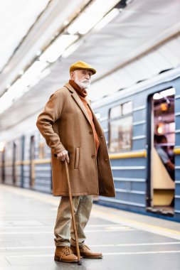 Başlık ve palto giymiş yaşlı bir adam bulanık arka planda metro istasyonunda duruyor.