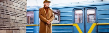 Güz giysileri içinde yaşlı bir adam metro peronunda duruyor arkasında tren, afiş