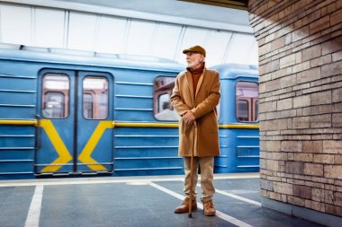 Güz kıyafetli yaşlı adam trenin yanındaki metro istasyonunda bastonla ayakta duruyor.