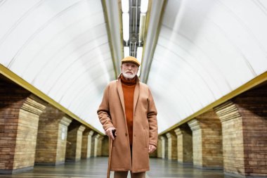 Son sınıf şapkalı ve sonbahar kıyafetli bir adam metro istasyonunda bastonla dikilirken kameraya bakıyor.
