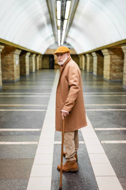 Güz ceketli ve şapkalı yaşlı adam elinde bastonla metro istasyonunda duruyor.