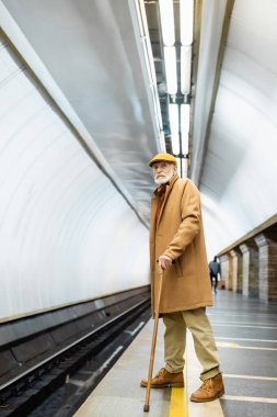 Sonbahar kıyafetli yaşlı bir adam elinde bastonla yeraltı platformunda duruyor.