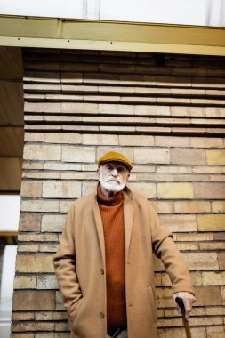 elderly man in autumn clothes standing near brick column at train station   clipart