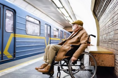 Tekerlekli sandalyedeki yaşlı adam, sonbahar kıyafetleri giyiyor, metro istasyonunda trenin yanında.