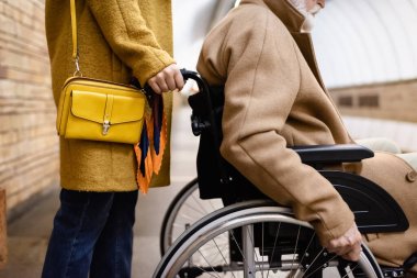 Metro istasyonunda yaşlı engelli bir adamın tekerlekli sandalyesini tutan kadın görüntüsü.