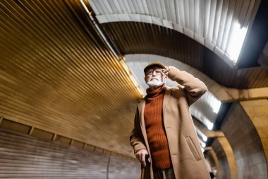 Sonbahar kıyafetleri içinde yaşlı bir adam metro platformunda dikilirken gözlüğe dokunuyor.