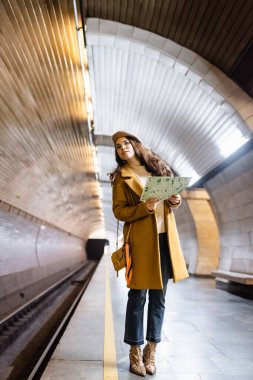 stylish woman in autumn clothes looking away while holding city map on underground platform clipart