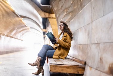 Şık sonbahar kıyafetleri içindeki genç kadın metro platformunda otururken elinde dergi tutuyor.