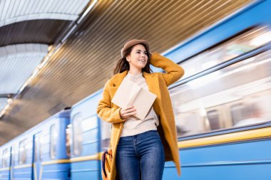 Sonbahar kıyafetleri içinde mutlu bir kadın kitap tutuyor ve metro treninin platforma gelişine bakıyor.