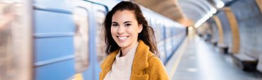 Bulanık tren ve pankartıyla yeraltı platformunda kameraya bakan mutlu genç kadın.