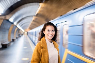 Güz ceketli mutlu kadın bulanık arka planda metro platformunda dikilirken kameraya gülümsüyor.