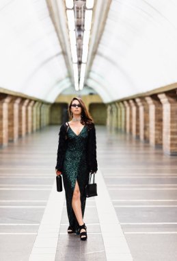 young elegant woman in long black dress and sunglasses standing on metro station with wine bottle and handbag clipart