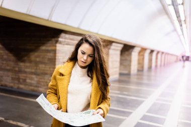 Güz ceketli genç kadın yeraltı istasyonunda şehir haritası okuyor.