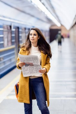 Güz kıyafetli genç bir kadın platformda bulanık metro treninin yanında gazete tutuyor.