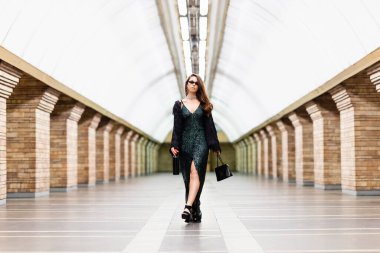 Uzun siyah elbiseli baştan çıkarıcı bir kadın elinde şarap şişesi ve çantasıyla metro istasyonunda yürüyor.