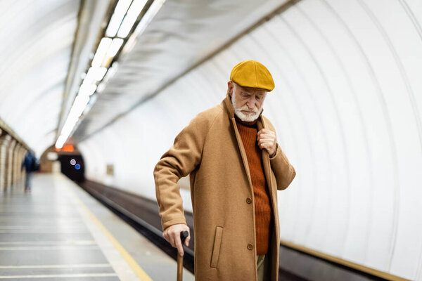 пожилой мужчина в шапке и осеннем пальто, стоящий с тростью на подземной платформе