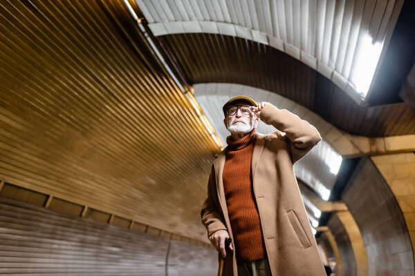 пожилой мужчина в осенней одежде трогает очки, стоя на платформе метро