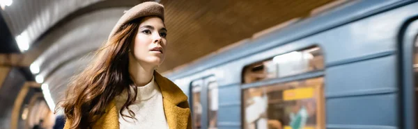年轻女子在秋天的贝雷帽和外套在地下铁车站的模糊的火车旁看着远方 一面横幅 — 图库照片