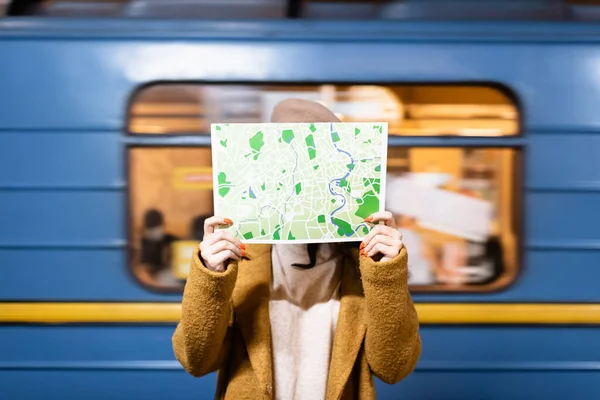 woman in autumn coat obscuring face with city map with blurred metro train on background