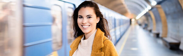 happy young woman looking at camera on underground platform with blurred train, banner