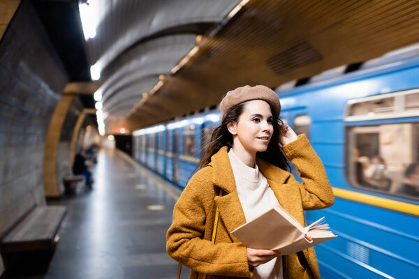 smiling woman in autumn outfit holding book while looking at blurred train on metro platform