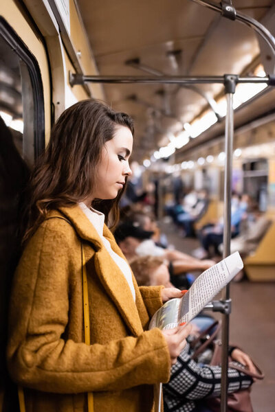 young woman in autumn coat reading newspaper while traveling in metro train