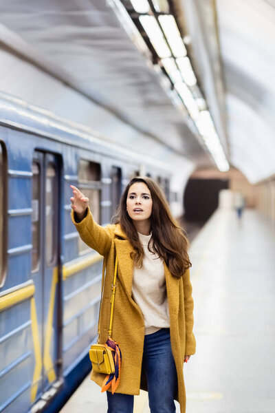 young woman in autumn outfit gesturing with outstretched hand near blurred train on metro platform 