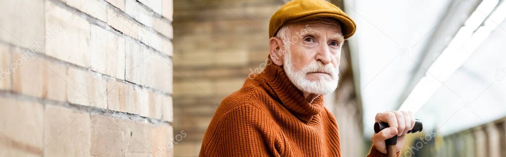 senior man in sweater and cap looking away while sitting on metro platform on blurred foreground, banner