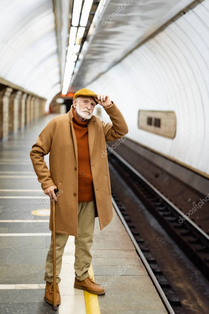 senior man in autumn outfit touching cap while standing on metro station platform