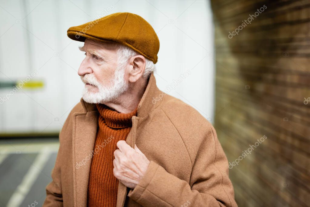 senior man in cap touching collar of autumn coat while standing at metro station and looking away