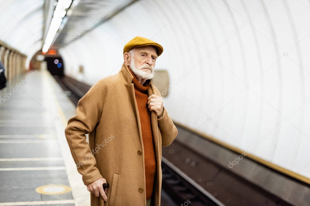 senior man in autumn outfit touching collar of coat while looking away on metro platform