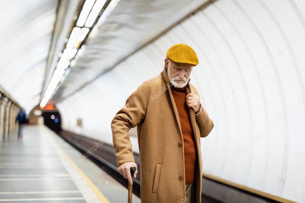 aged man in cap and autumn coat standing with walking stick on underground platform