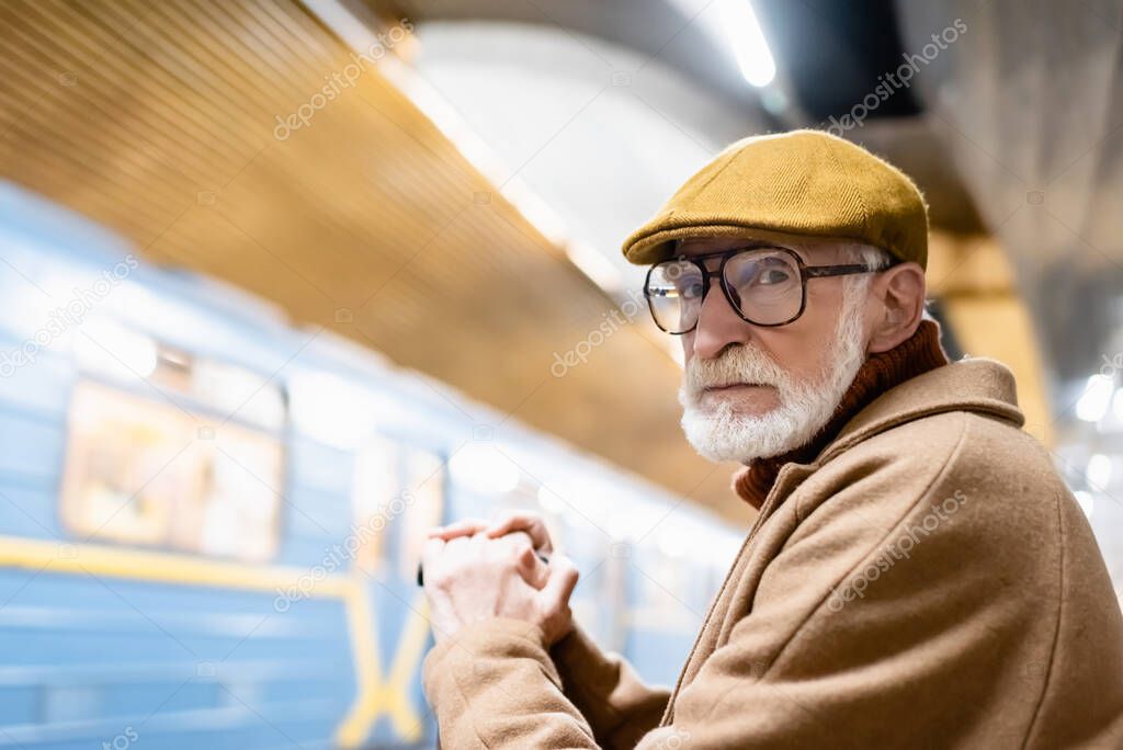 senior man in autumn clothes and eyeglasses looking at camera on metro platform with blurred train